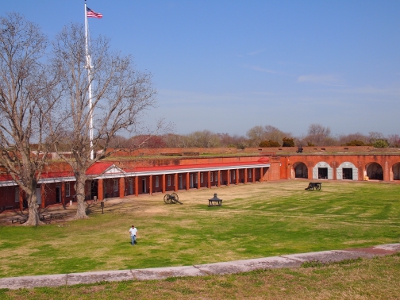 [Looking onto the fort grounds inside the fort while standing atop the fort wall. Tall flag pole stands above two large leafless tress inside the fort grounds. Several cannons on the grass.]